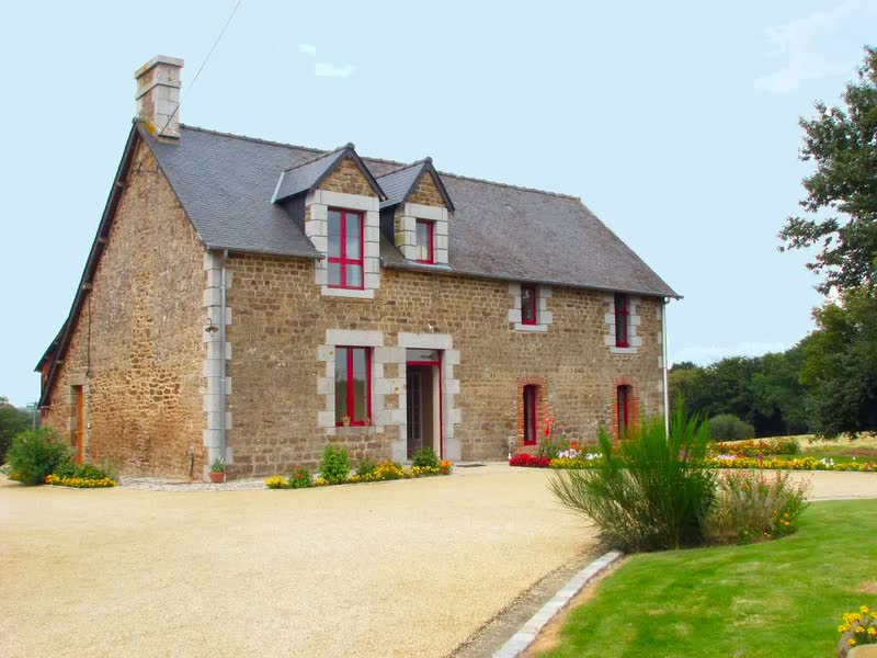 La maison en pierre du bocage mayennais sous un ciel bleu, cour gravillonnée et massifs fleuris